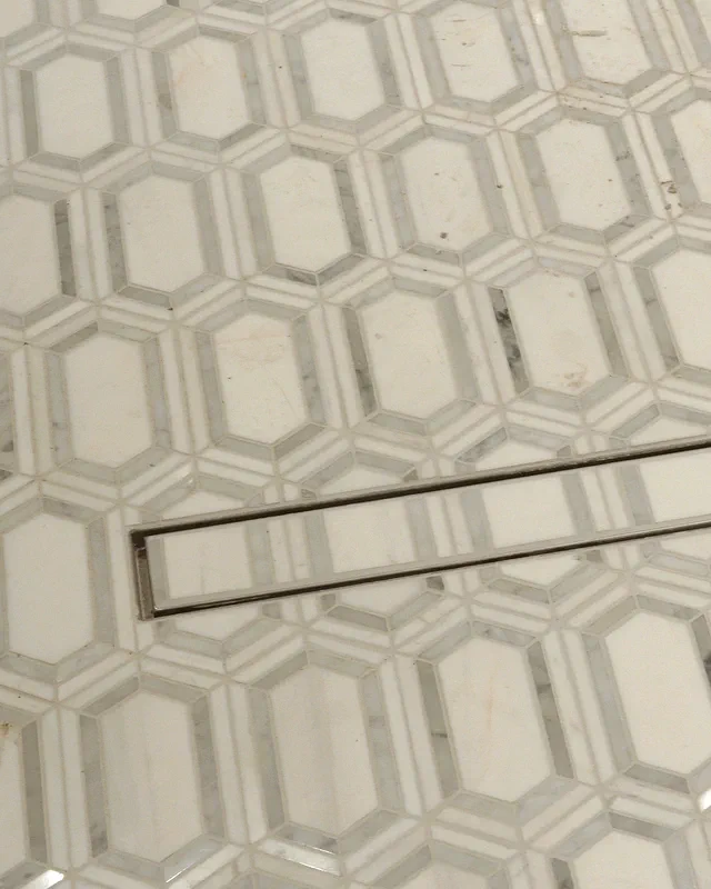 A long, narrow stainless shower drain set into white hexagon floor tile.