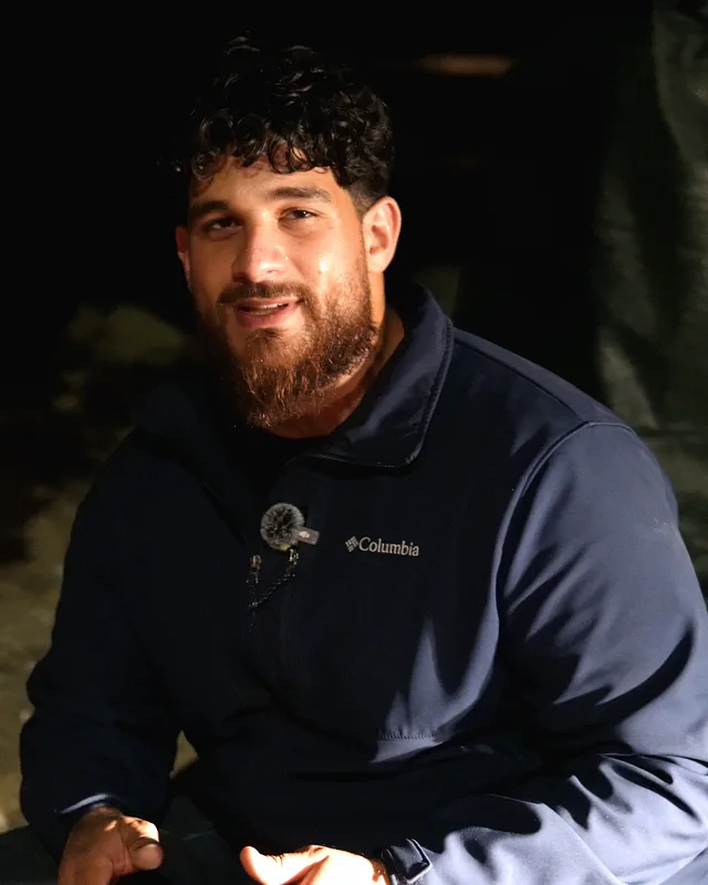 The owner of H&S Plumbing crouched outside at night, talking to the camera beside a water meter box.